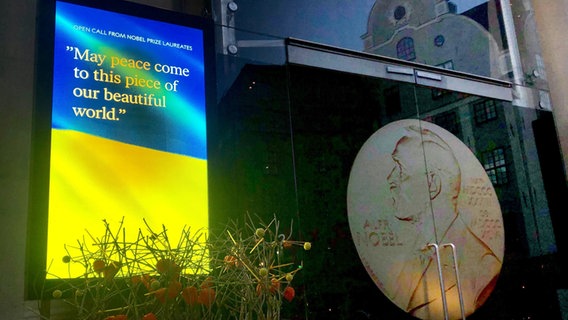 Auf einem Bildschirm vor dem Nobelpreismuseum in Stockholm ist eine Ukraine-Flagge zu sehen mit dem Schriftzug "May peace come to this piece of our beautiful world" © dpa-Bildfunk Foto: Steffen Trumpf/dpa