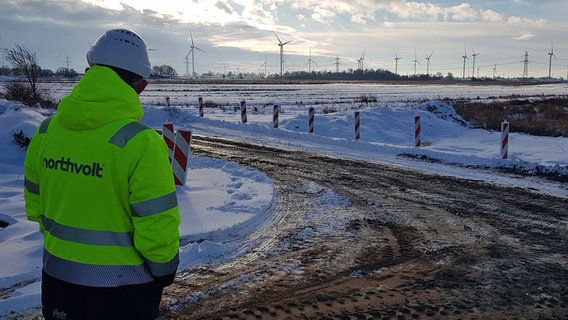 Ein Bauarbeiter mit einer Jacke mit der Aufschrift "Northvolt" steht auf einer schneebedeckten Baustelle, im Hintergrund sind Windräder zu sehen. © NDR Foto: Lukas Knauer