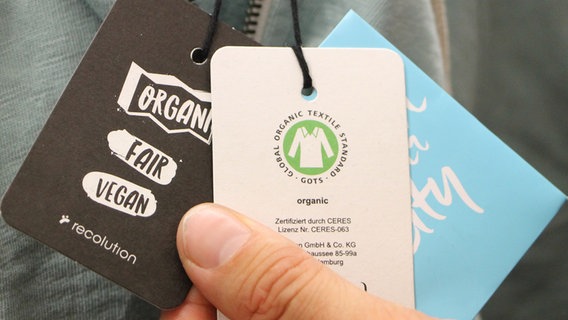 Das GOTS-Schild eines Organic Fashion Labels wird in einer Hand gehalten. © NDR Foto: Micheal Latz