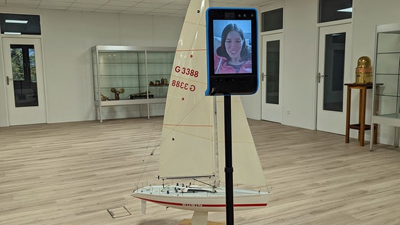 Auf einem auf einem Segway montierten Smartphone, das in einem Raum mit einem Segelboot im Hintergrund steht, ist Elke Paatz, Inklusionsbeauftragte des Deutschen Seglerverbands, zu sehen. © NDR Foto: Astrid Kühn