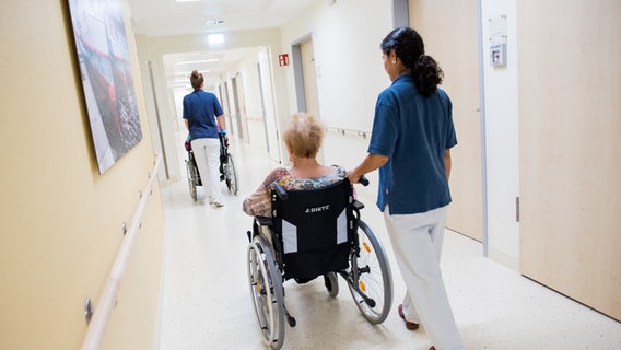 Zwei Pflegekräfte schieben jeweils einen Rollstuhl mit einer Person einen Gang entlang © picture alliance/dpa Daniel Bockwoldt Foto: Daniel Bockwoldt