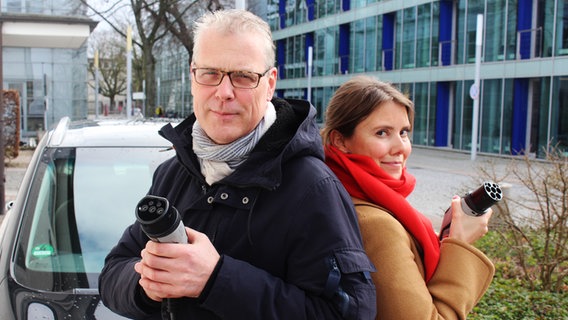 Nicolas Lieven und Susanne Tappe stehen Rücken an Rücken mit einem Ladekabel für Elektrofahrzeuge in der Hand. © NDR Foto: Jürgen Webermann