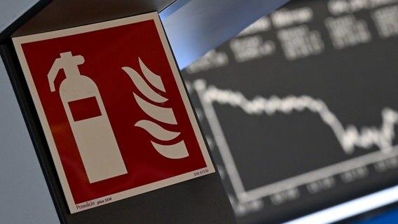 Ein Hinweisschild für einen Feuerlöscher ist an einem Durchgang der Frankfurter Wertpapierbörse angebracht, während die Anzeigetafel mit der Dax-Kurve fallende Kurse anzeigt. © picture alliance Foto: Arne Dedert