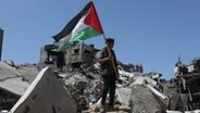 Ein Palästinenser hält die palästinensische Fahne in der Hand, während er auf Trümmern nach israelischem Beschuss in Chan Junis (Süd-Gaza) steht (Archivbild vom 13.4.24) © dpa Foto: Abed Rahim Khatib