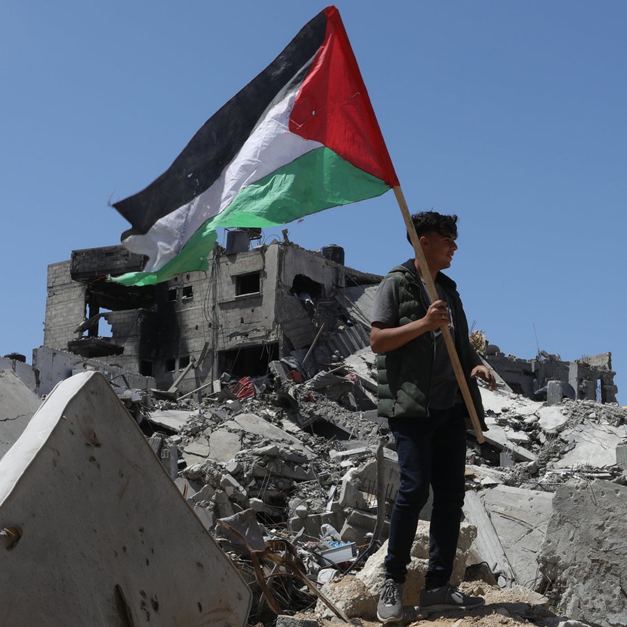 Ein Palästinenser hält die palästinensische Fahne in der Hand, während er auf Trümmern nach israelischem Beschuss in Chan Junis (Süd-Gaza) steht (Archivbild vom 13.4.24) © dpa Foto: Abed Rahim Khatib