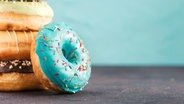 Ein Stapel von Donuts mit einem Donut mit blauer Glasur und bunten Streuseln im Vordergrund. © picture alliances Foto: Olga Sergeeva