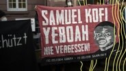 Ein Demonstrant zeigt ein übergroßes Plakat mit dem Namen und einem Abbild eines Mannes in Erinnerung an den bei einem Brandanschlag auf eine Asylunterkunft verstorbenen Samuel Kofi Yeboah. © picture alliance/dpa Foto: Thomas Frey