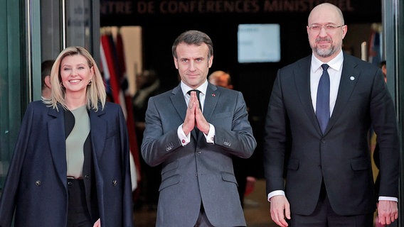 Die ukrainische First Lady Olena Zelenska (l.), der französische Präsident Emmanuel Macron (M.) und der ukrainische Premierminister Denys Shmyhal stehen vor einem Gebäude nebeneinander. © dpa bildfunk/AP Foto: Lewis Joly
