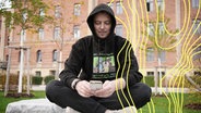Hacker Benjamin Gnahm sitzt mit einem Kapuzenpulli im Freien vor einem Gebäude und tippt auf seinem Smartphone. © BR Foto: Olaf Bitterhoff