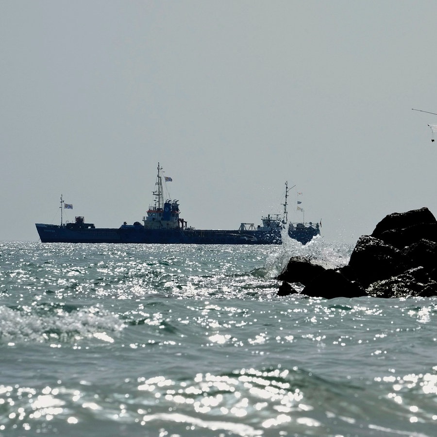 Zwei Männer angeln am Meer, während im Hintergrund ein mit 240 Tonnen Konserven und Hilfsgütern für den Gazastreifen beladenes Schiff in den Gewässern vor dem zypriotischen Hafen von Larnaca fährt. © AP/dpa Foto: Petros Karadjias