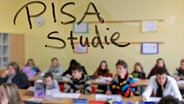 Die Wörter "PISA Studie" stehen auf einer Glasscheibe in einer Schulklasse, während Schüler den Unterricht verfolgen. © dpa Foto: Julian Stratenschulte
