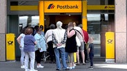 Kunden stehen in einer Warteschlange vor einer Filiale einer Postbank. © picture alliance 