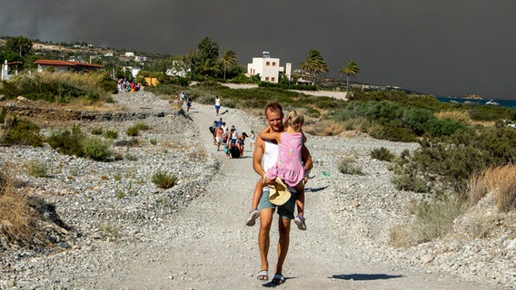 Ein Mann trägt ein Kind, als sie ein Waldbrandgebiet auf Rhodos (Griechenland) verlassen. © InTime News/AP/dpa Foto: Lefteris Damianidis