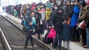 Viele Menschen stehen auf einem Bahnsteig dicht aneinander gedrängt, im Vordergrund läuft eine Frau mit einem Kind auf dem Arm über Bahnschienen © picture alliance / dmg media Licensing | Jamie Wiseman Foto: Jamie Wiseman