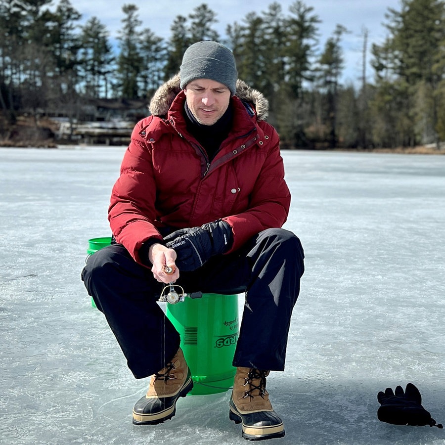 Ingo Zamperoni sitzt in den USA zusammen mit Coach auf einem zugefrorenen See und angelt in einem ins Eis geschlagenen Loch. © NDR Foto: Martin Kobold