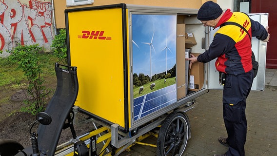 Ein Paketzusteller steht vor einem Hauseingang neben einem zu einem Zustellwagen umgebauten Dreirad: In einer großen, an der Seite geöffneten Box sind viele Pakete zu sehen. © NDR Foto: Michael Latz