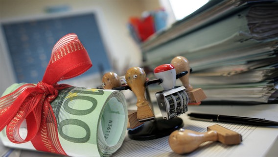 Gerolltes Geldbündel mit Schleife vor einem Behördenschreibtisch (Montage) © picture-alliance/ ZB  --- fotolia Foto: Patrick Pleul --- Joachim Wendler