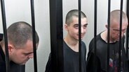 Zwei britische Staatsbürger (links und rechts) und ein Marokkaner sitzen hinter Gittern in einem Gerichtssaal in Donezk, in dem von der Regierung der Volksrepublik Donezk kontrollierten Gebiet im Osten der Ukraine. © Uncredited/AP/dpa 