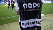 Am Rand eines Fußballspielfeld steht ein Mann, auf dessen Rücken "Dopingkontrolle - NADA - für sauberen Sport" steht. © dpa picture alliance Foto: Hansjürgen Britsch