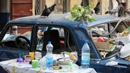 Tauben sitzen auf einem zerstörten Auto und suchen nach Essen, davor steht ein Tisch mit Geschirr und Essensresten. © Anadolu Agency Foto: Leon Klein