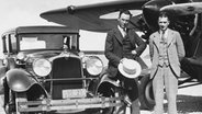 Gründer Howard Hughes der "Pacific Aero Products Company" steht neben dem Kunstflieger Roscoe Turner mit einem Auto und einem Flugzeug aus seiner Herstellerfirma. © Imago/UIG 
