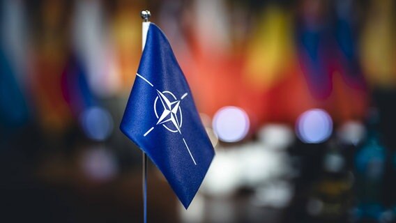 Eine kleine Flagge mit dem Logo der NATO steht auf einem Tisch. © IMAGO/photothek Foto: Xander Heinl