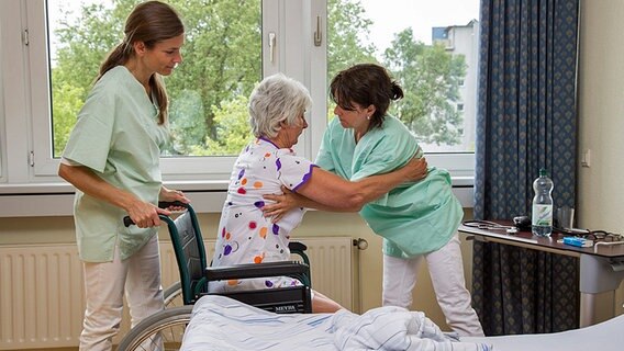 Zwei Pflegerinnen helfen in einem Zimmer einer Patientin aus dem Rollstuhl ins Bett. © dpa bildfunk Foto: AOK-Mediendienst