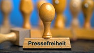 Ein Holzstempel mit der Aufschrift "Pressefreiheit" auf einer Schieferplatte mit weiteren Stempeln unscharf dahinter. Foto mit Composing vor einem blauen Hintergrund. © picture alliance / SULUPRESS.DE Foto: Torsten Sukrow