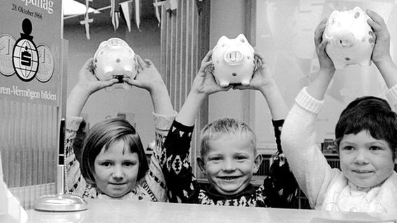 Drei Kinder halten am 28.10.1966, dem Weltspartag, stolz ihre Sparschweine vor einem Sparkassenschalter in die Höhe. © picture-alliance / dpa 