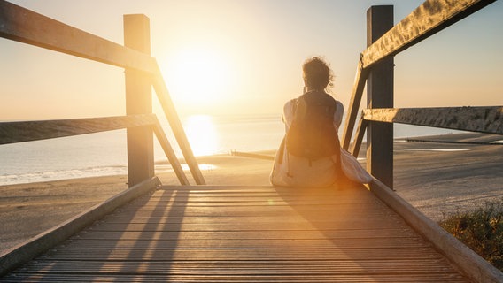 Eine Person sitzt auf einer Holztreppe am Strand und schaut in die untergehende Sonne. © photocase.de Foto: rclassen