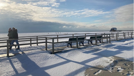 Sankt Peter Ording: Passanten gehen auf einem Steg über schneebedeckte Dünen spazieren. © Christiane Bosch/dpa 