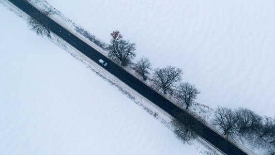 Eine Luftaufnahme zeigt ein Auto, das auf einer geräumten Straße durch eine verschneite Winterlandschaft fährt. © picture alliance/dpa | Jens Büttner 
