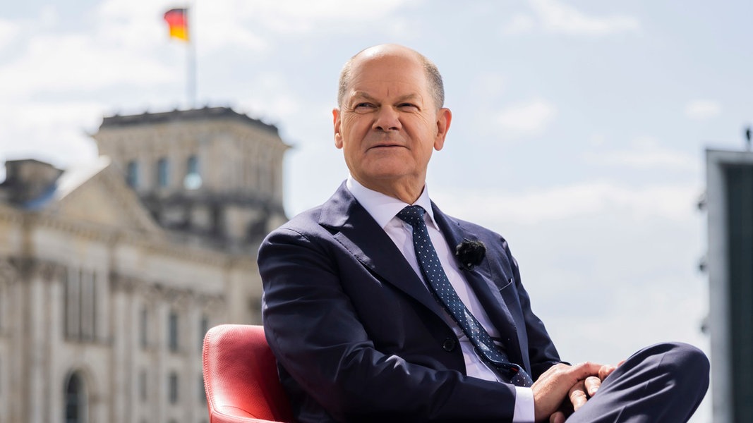 Bundeskanzler Olaf Scholz sitzt vor einem Interview in einem Sessel, im Hintergrund das Reichstagsgebäude.