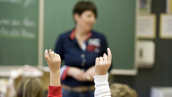 Unterricht in einer Grundschulklasse: Schülerinnen melden sich mit Handzeichen, vor der Tafel steht die Lehrerin. © picture alliance / dpa Foto: Ulrich Baumgarten