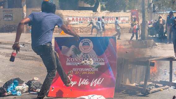 Ein wütender Protestant tritt auf ein Plakat ein. Dahinter brennt es. © dpa picture alliance Foto: Sabin Johnson