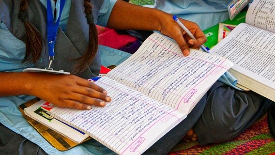 Ein Schulkind sitzt in der Schule auf dem Boden mit einem Schulheft und einem Schulbuch. © picture alliance Foto: Lena Iliopoulou