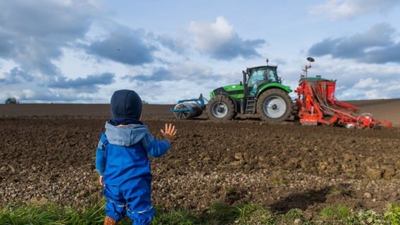 Ein kleines Kind steht am Rand eines Ackers und winkt einem Traktor zu. © picture-alliance/dpa Foto: Benjamin Nolte