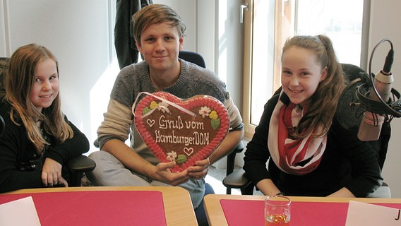 Zwei Kinder sitzen im Hörfunkstudio neben dem Moderator, der ein großes Lebkuchenherz in der Hand hält © NDR Foto: Katharina Mahrenholtz