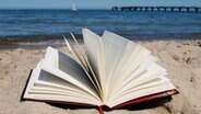 Ein Buch liegt aufgeschlagen am Strand, im Hintergrund die Ostsee. © Katharina Mahrenholtz Foto: Katharina Mahrenholtz