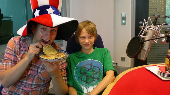 Im Studio beißt der Moderator, der einen großen USA-Hut trägt, von einem Pancake ab, neben ihm sitzt ein Junge. © NDR Foto: Katharina Mahrenholtz