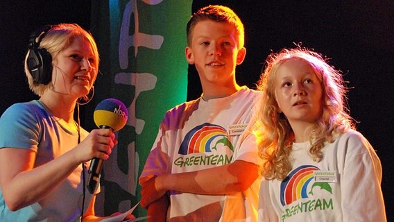 Eine Moderatorin und zwei Kinder stehen auf eine Bühne und richten ihren Blick nach links. © NDR Foto: Florian Jakobsen