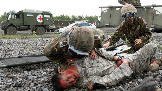 Ein Soldat liegt verletzt am Boden. Über ihn gebeugt zwei Sanitäter der Bundeswehr. Im Hintergrund Sanitätsfahrzeuge mit Rotem Kreuz. © Bundeswehr/Bienert Foto: Bienert