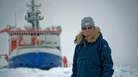 Markus Rex steht warm angezogen am Nordpo. Im Hintergrund ist das Forschungsschiff Polarstern zu sehen. © Markus Rex 