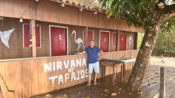Ein Mann mit blauem Oberteil steht vor einer Holzkonstruktion, darauf steht: Nirvana do Tapajos (Pedrinho) © NDR Foto: Tom Noga