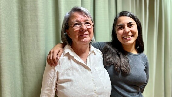 Eine ältere Frau steht vor einem grünen Vorhang, eine jüngere Frau mit dunklen langen Haaren umarmt diese lächelnd © NDR Foto: Max-Marian Unger