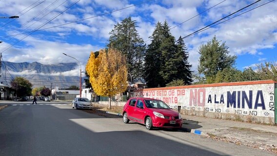 Ein rotes Auto steht auf einer Straße, eine Mauer trägt daneben die Aufschrift "no a la mina" und dahinter ein Bergpanorama und einige Bäume © NDR Foto: Max-Marian Unger
