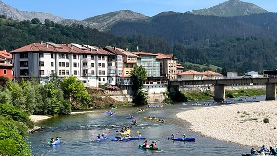 Menschen paddeln in Booten auf einem Fluss in Asturien, im Hintergrund eine Stadt, eine Brück und Berge © NDR Foto: Carsten Vick