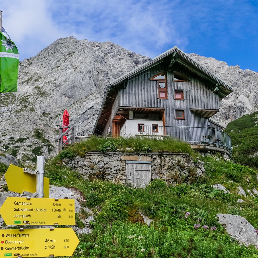 Eine Bergsteigerhütte in den Alpen, davor gelbe Schilder und eine grüne Flagge, dahinter die Alpen © NDR Foto: Jörg Wunram