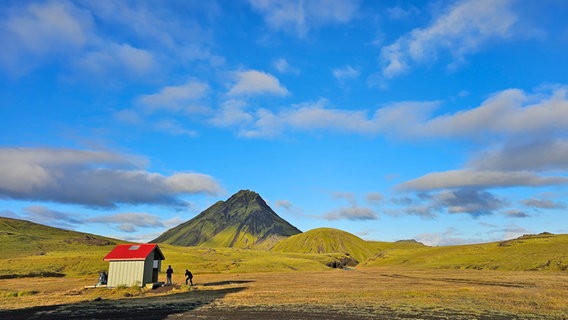 Eine winzige Holzhütte mit rotem Dach auf einer isländischen vulkanischen Ebene, davor stehhen zwei Personen © NDR Foto: Merle Schaack und Michael Richmann
