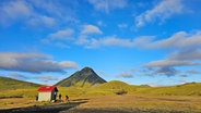Eine winzige Holzhütte mit rotem Dach auf einer isländischen vulkanischen Ebene, davor stehhen zwei Personen © NDR Foto: Merle Schaack und Michael Richmann